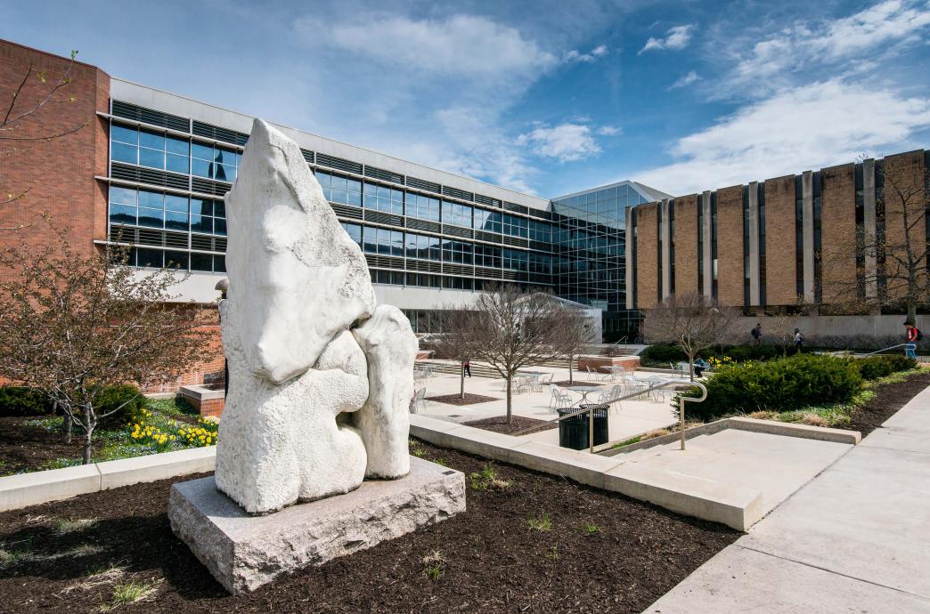 A large, light gray, stone, abstract sculpture stands in front of a campus building.
