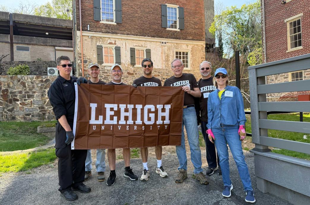 Alumni stand holding a brown Lehigh flag outside of stone buildings.