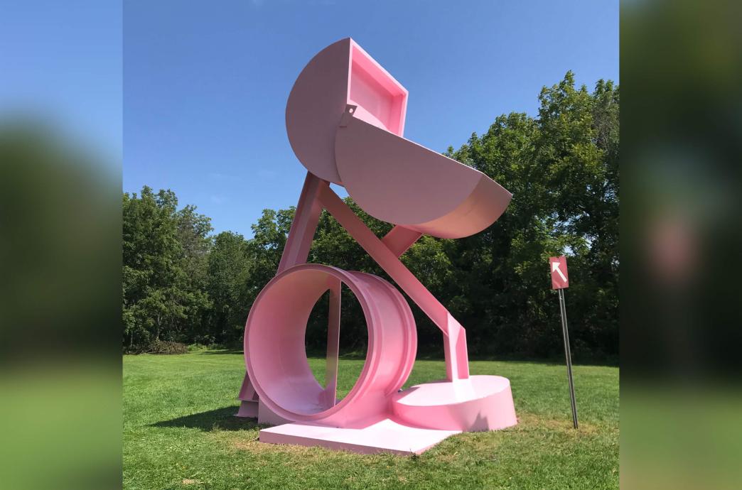 A refurbished, freshly painted, pink sculpture stands in a field during warmer months, with bright green grass and lush trees in the distance. 