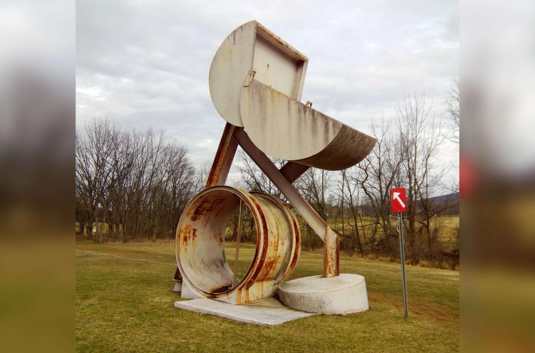 A large rusty sculpture stands in a field during cooler months, with bare trees in the distance. 