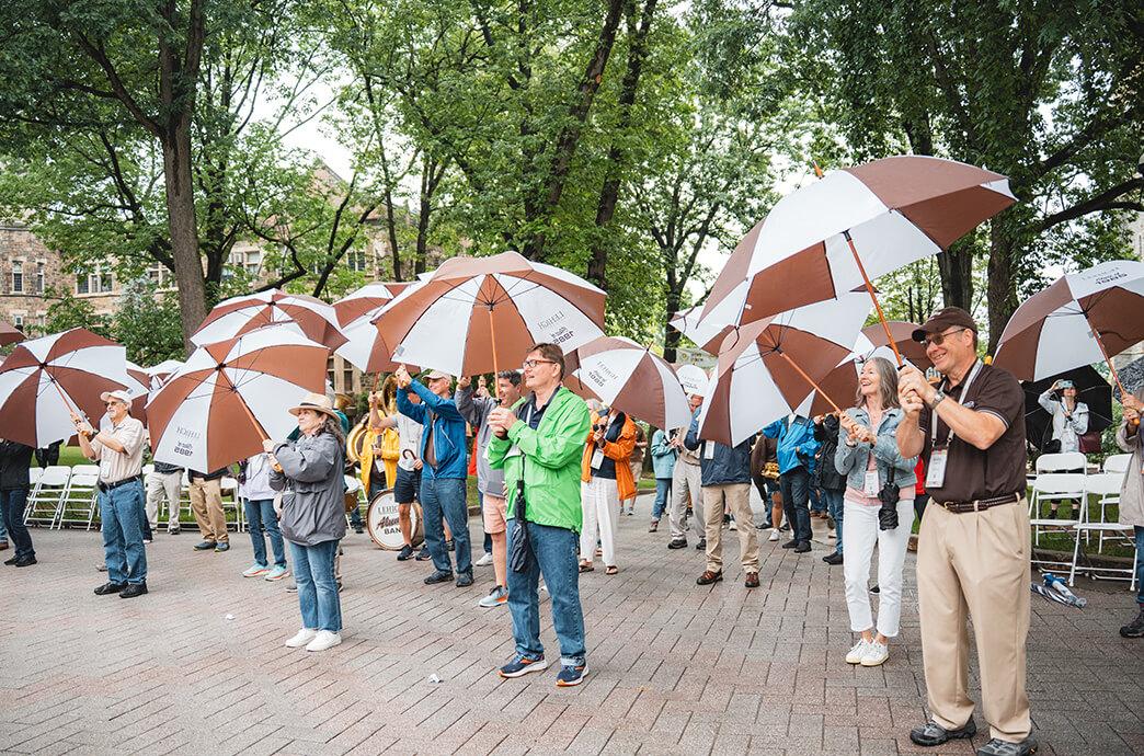  A group of smiling alumni stand outdoors under large brown and white umbrellas. 