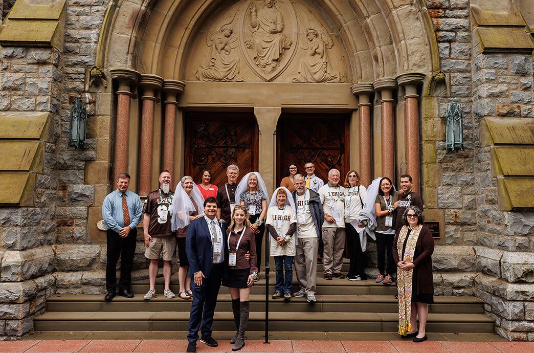  A group of people, some wearing wedding veils, pose for a photo on the steps of a stone church with ornate wooden doors.
