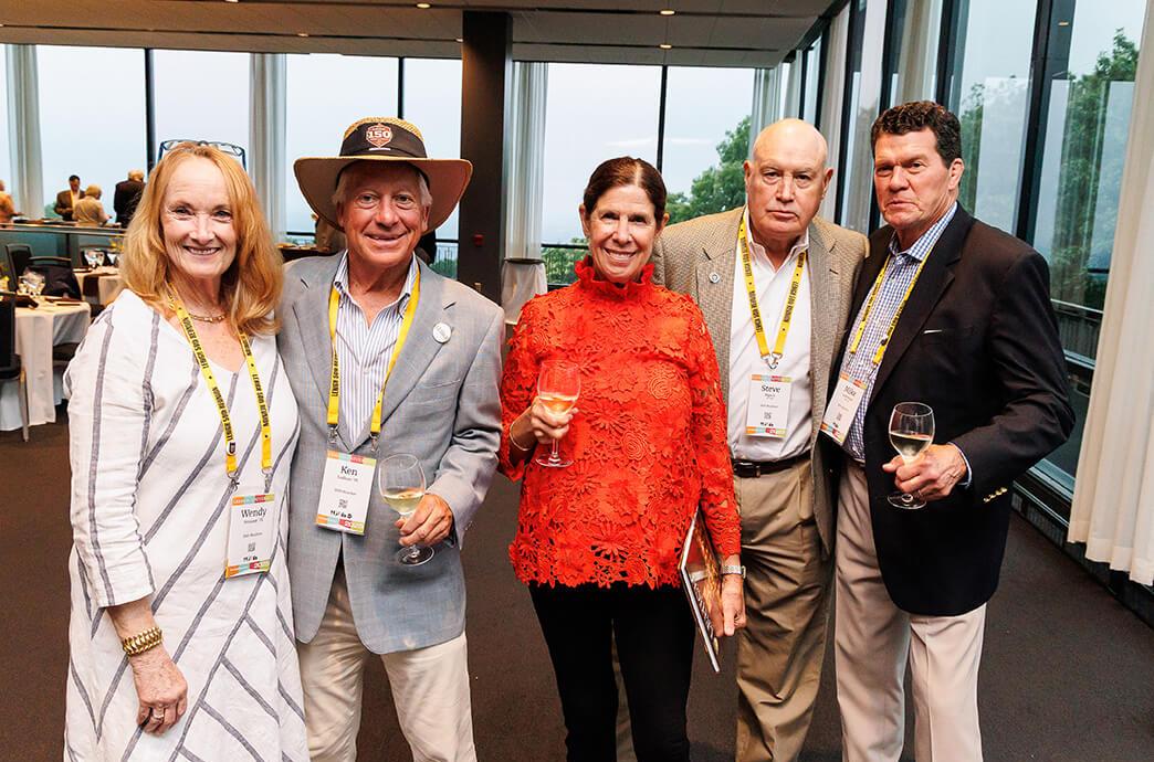 A group of five people stand together in a modern venue, each holding a glass of drink, dressed in semi-formal attire.