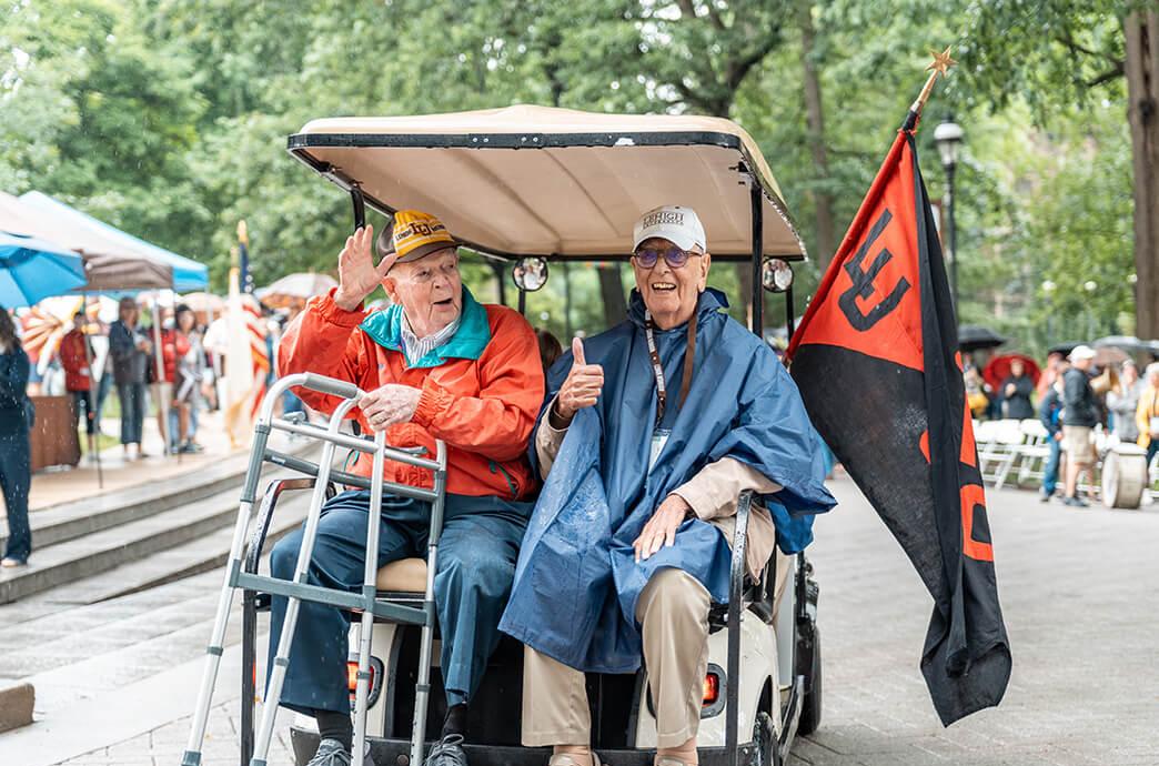 Two elderly men in a golf cart, one waves while holding a walker, and the other gives a thumbs up, surrounded by a lively crowd.