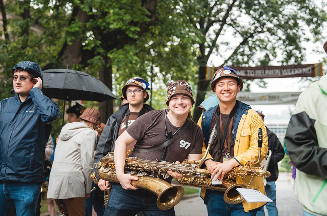 Two smiling individuals in colorful hats hold saxophones, surrounded by others in raincoats and umbrellas at an outdoor event