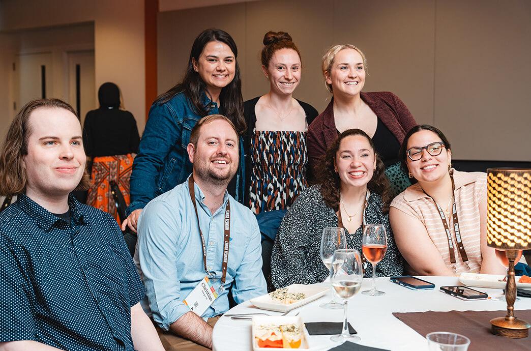 A group of seven people smiling around a table at a social event. The setting is warm and friendly.