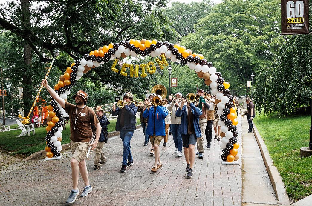  A Marching band under a balloon arch reading "Lehigh" in festive colors of black, white, and gold.