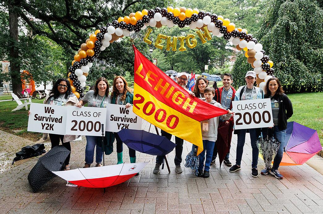 A group of smiling alumni stand under a balloon arch holding signs, including “We Survived Y2K” and “Class of 2000.” 