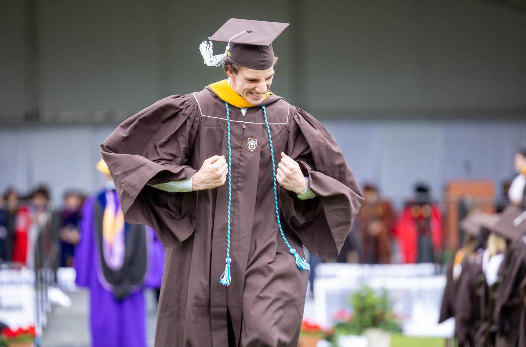 A male graduate wearing a brown cap and gown points flexes his two arms with enthusiasm as he walks back to his seat.