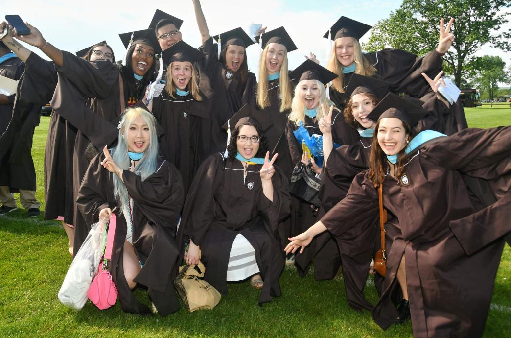 A group of female graduates squat and huddle for a photo, making enthusiastic faces and motions for the camera while holding their personal items and purses.