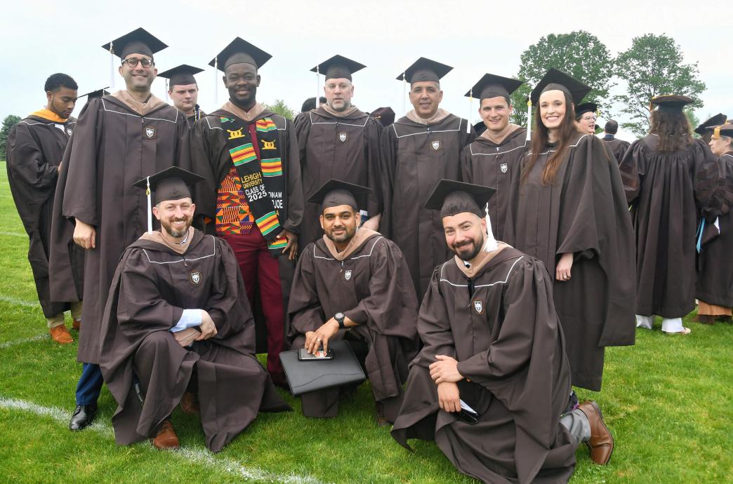 A group of graduates in brown caps and gowns pose for a photo on the field of Goodman Stadium, with some squatting in front of the group.