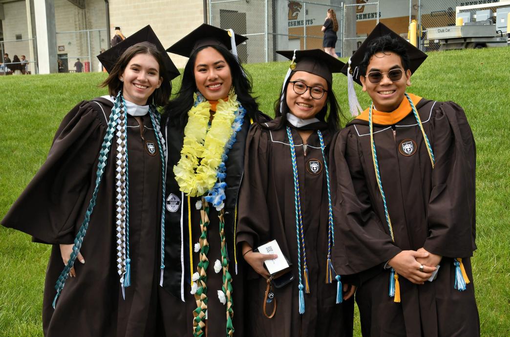 Four graduates in brown caps and gowns pose for a photo with big smiles, one wearing a lei.