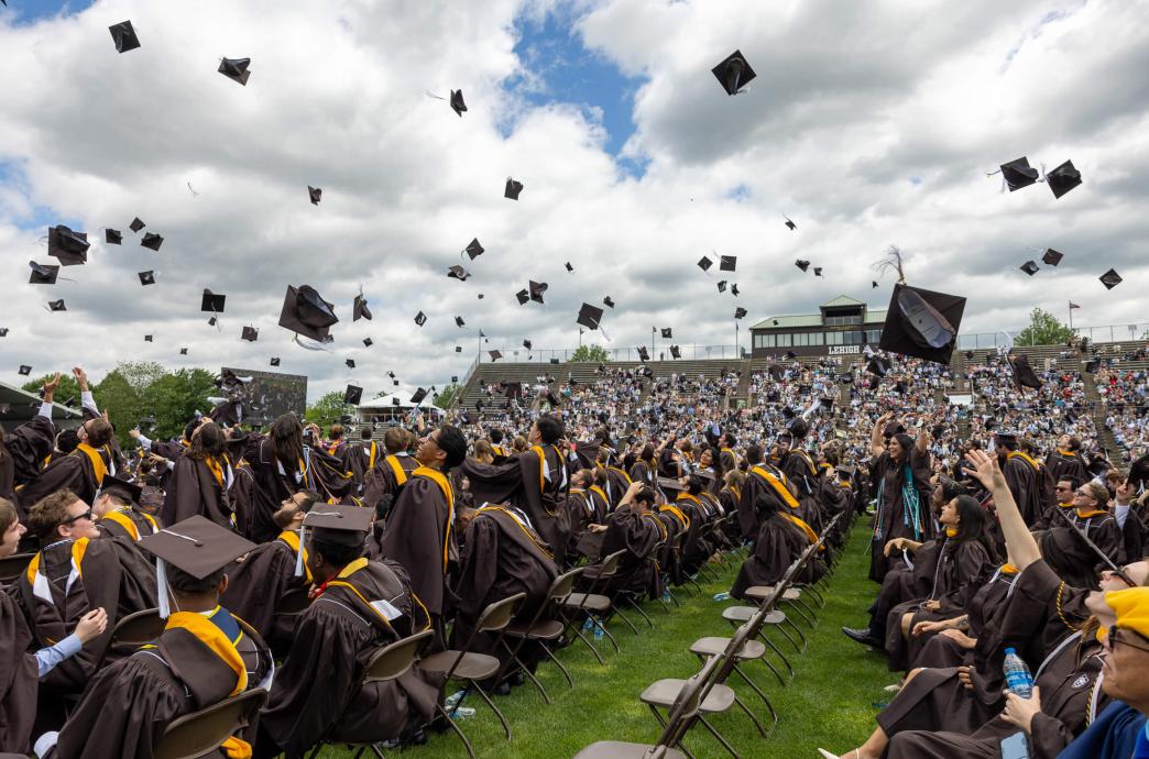 A wide shot shows a full crowd of graduates with caps tossed in the air with clouds visible in the blue sky.