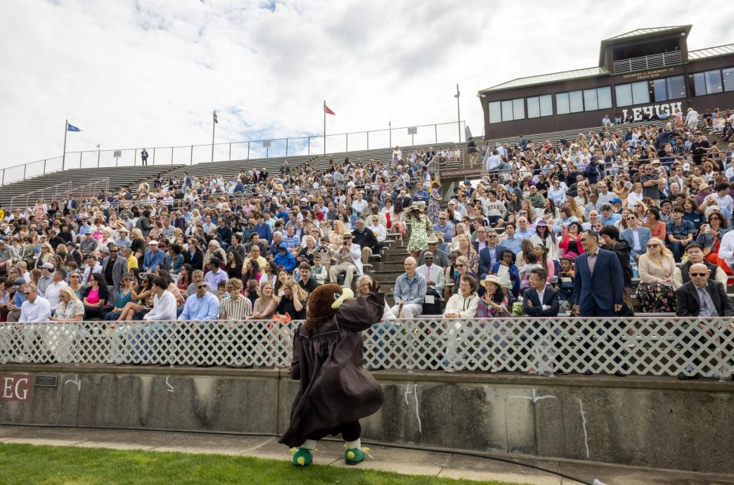 A performer in a mountain hawk costume and brown graduation gown hypes up an audience of families in the Goodman Stadium stands.