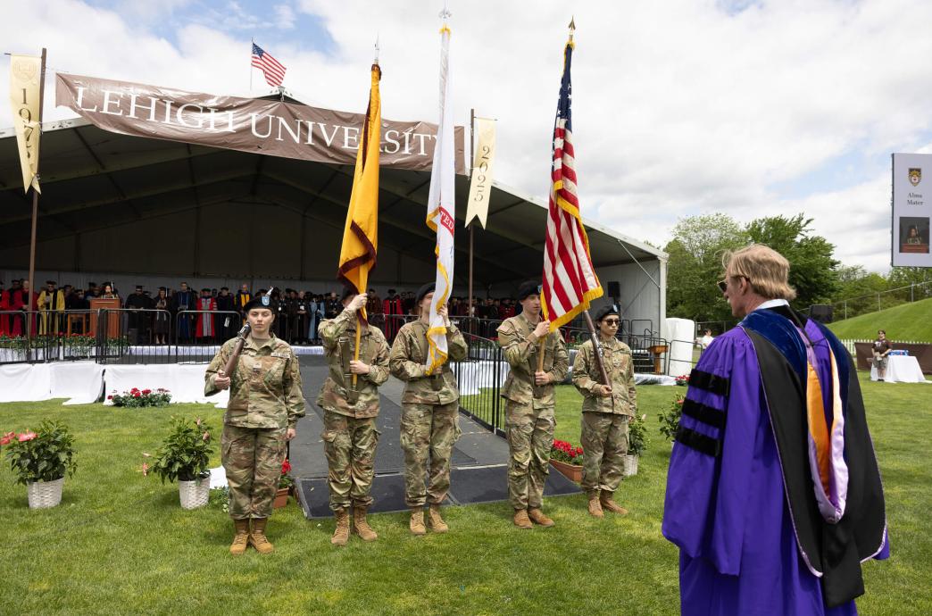 Five people in military fatigues hold flags in front of a professor in purple regalia.