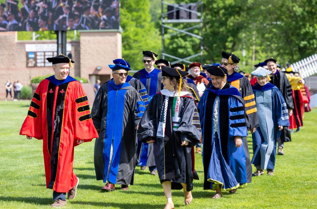 Professors and administration in various colors of regalia walk down the field in 2 rows.