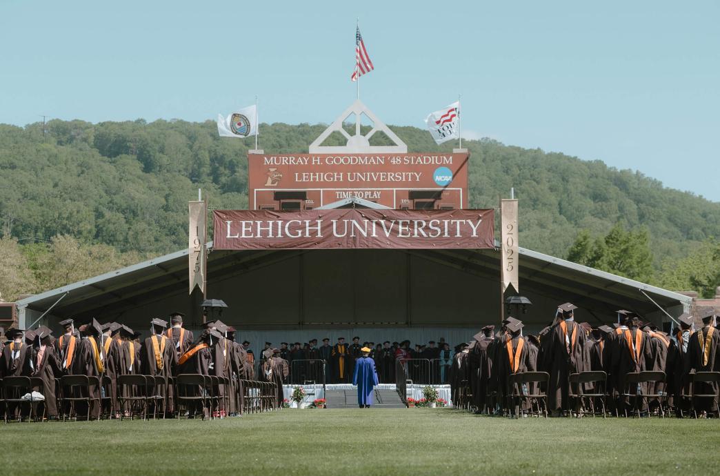 Rows of graduates in brown ropes stand at their seats at Goodman Stadium in front of a stage with a Lehigh University banner hanging above it.
