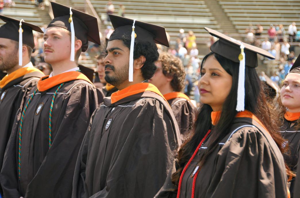 The camera focuses on 3 students wearing brown caps and gowns in the assembly of graduates during their ceremony.
