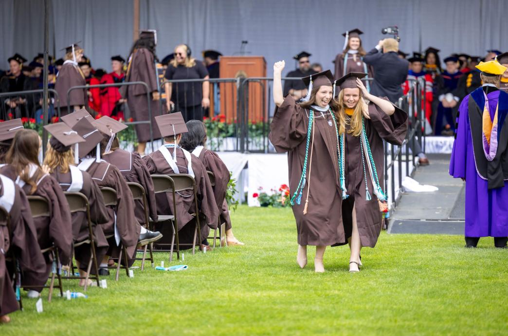 Returning from the stage, 2 female graduates wearing brown caps and gowns embrace side-by-side and celebrate walking between the aisle of fellow graduates.