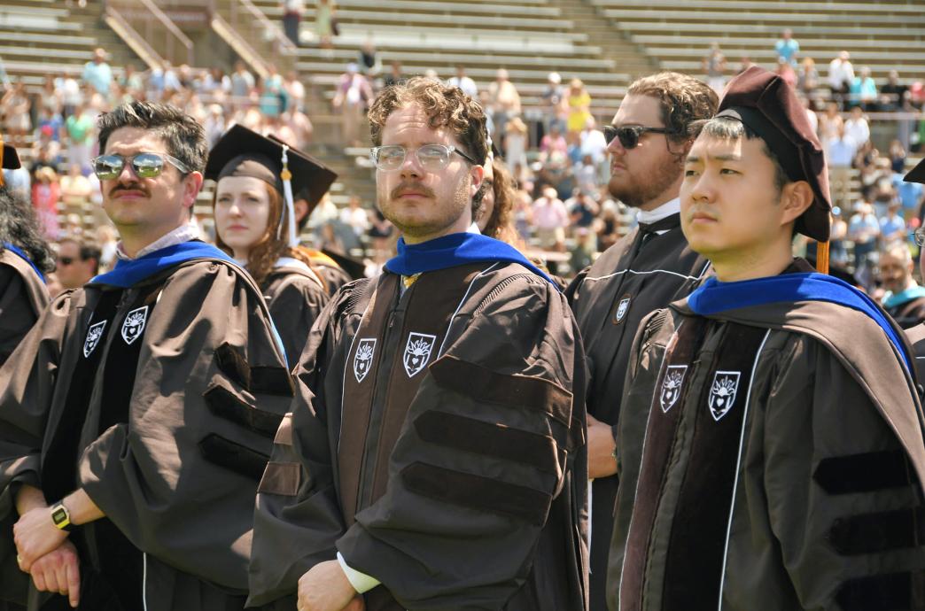The camera focuses on 3 doctoral graduates wearing brown caps and gowns in the assembly of graduates during their ceremony.