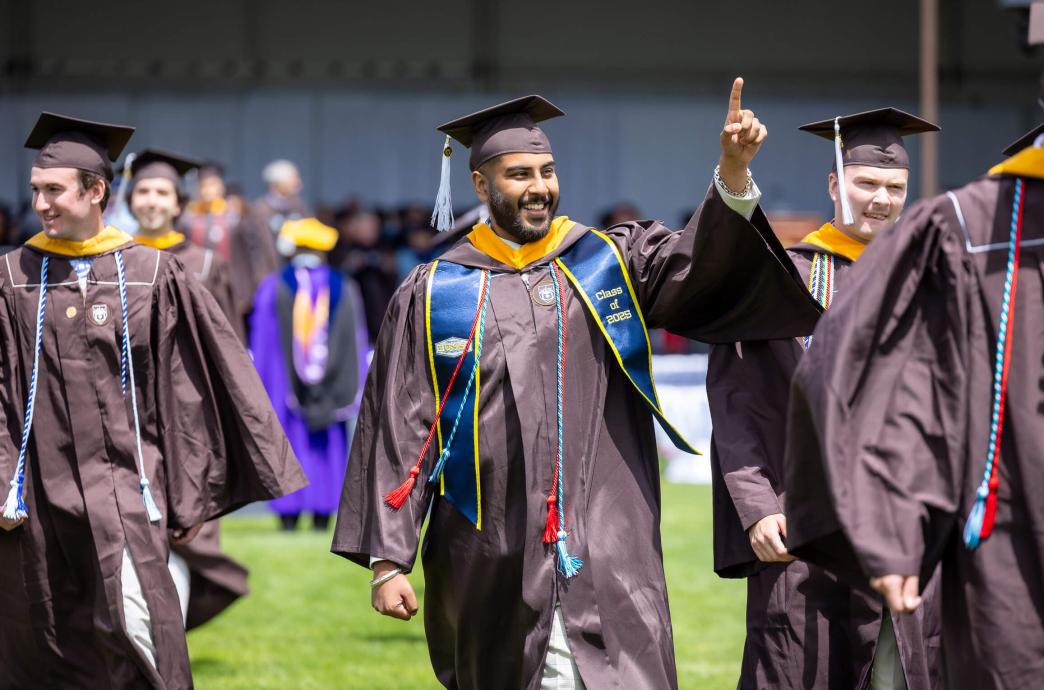 Returning from the stage, a male graduate wearing a brown cap and gown points with his hand in the sky to crowd as he walks among fellow graduates.