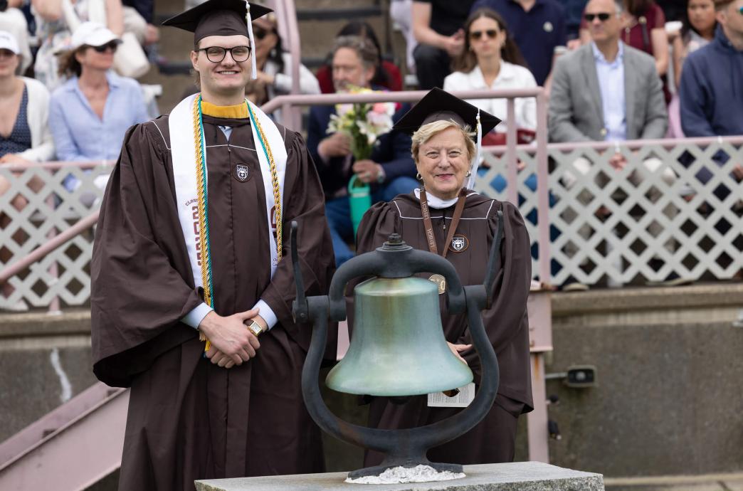Both wearing brown graduation caps and gowns, a tall male student and an alumna from the class of 1975 pose for a photo with a large ceremonial bell.
