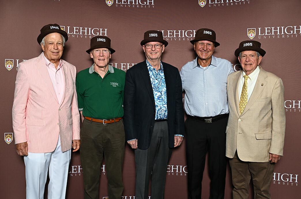 Five older men stand smiling in front of a Lehigh University backdrop, wearing hats labeled "1965."