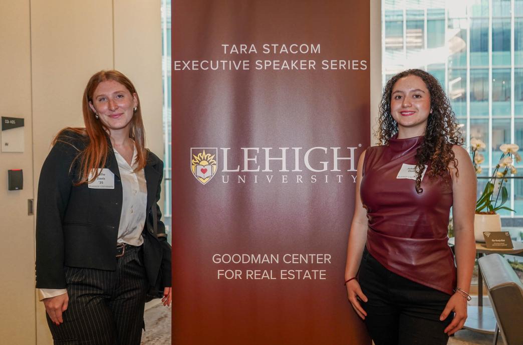 Two women dressed professionally flank a tall brown sign reading “Tara Stacom Executive Speaker Series, Lehigh University, Goodman Center for Real Estate”
