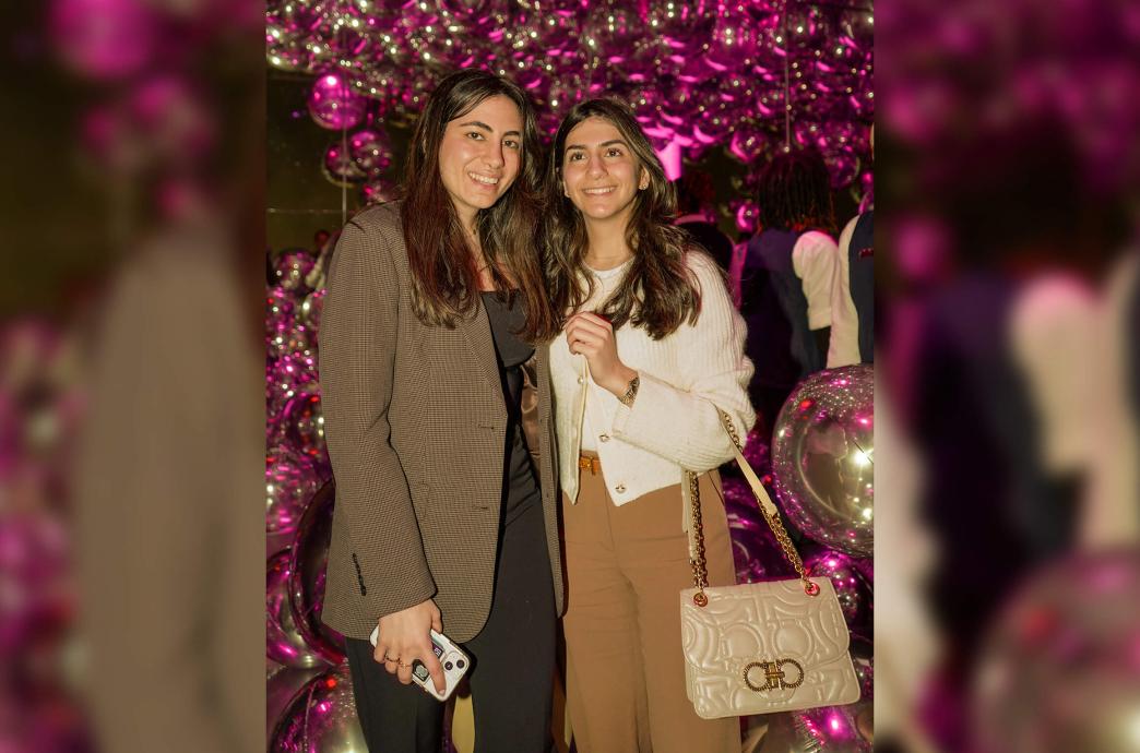 Two women, one carrying a cream-colored purse on her forearm, pose in a room filled with fuschia light illuminated balloons.