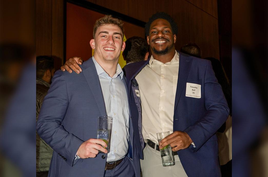 Two men holding drink glasses smile and pose, one with his hand on the shoulder of the other.