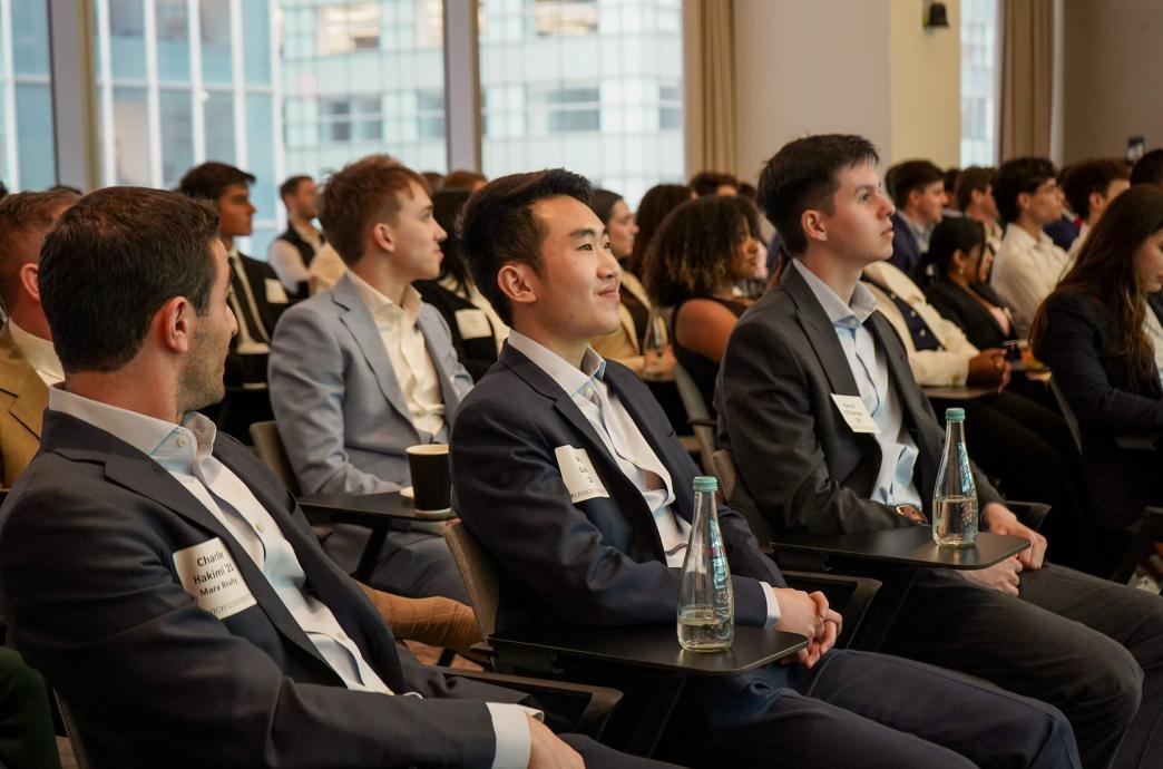 A room of event attendees sit in a conference room, all wearing suits and nametags, with a focus on 3 men in the front row.