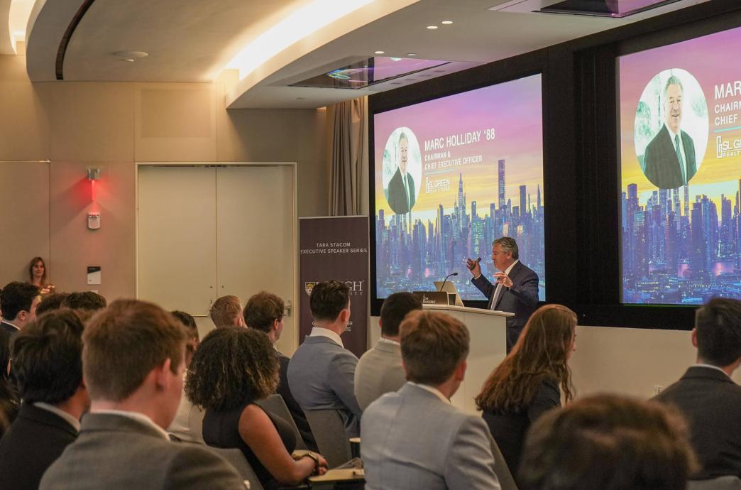 Event attendees seated facing a man speaking at a podium with large screens behind him, reading Mark Holliday ’88, chairman & executive officer”