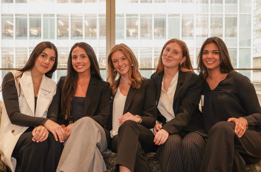 Five women in professional attire sit on a couch, side by side, smiling with their legs crossed.