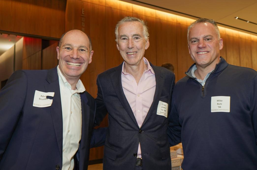 Three men dressed in business casual attire and wearing name tags, put their arms around each other and smile for a photo.