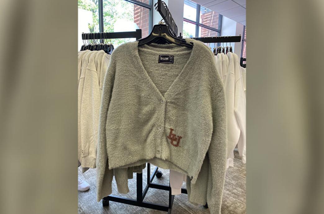 A cropped gray cardigan with the letters “LU” emblazoned on the bottom corner hangs on a clothing rack in a store.