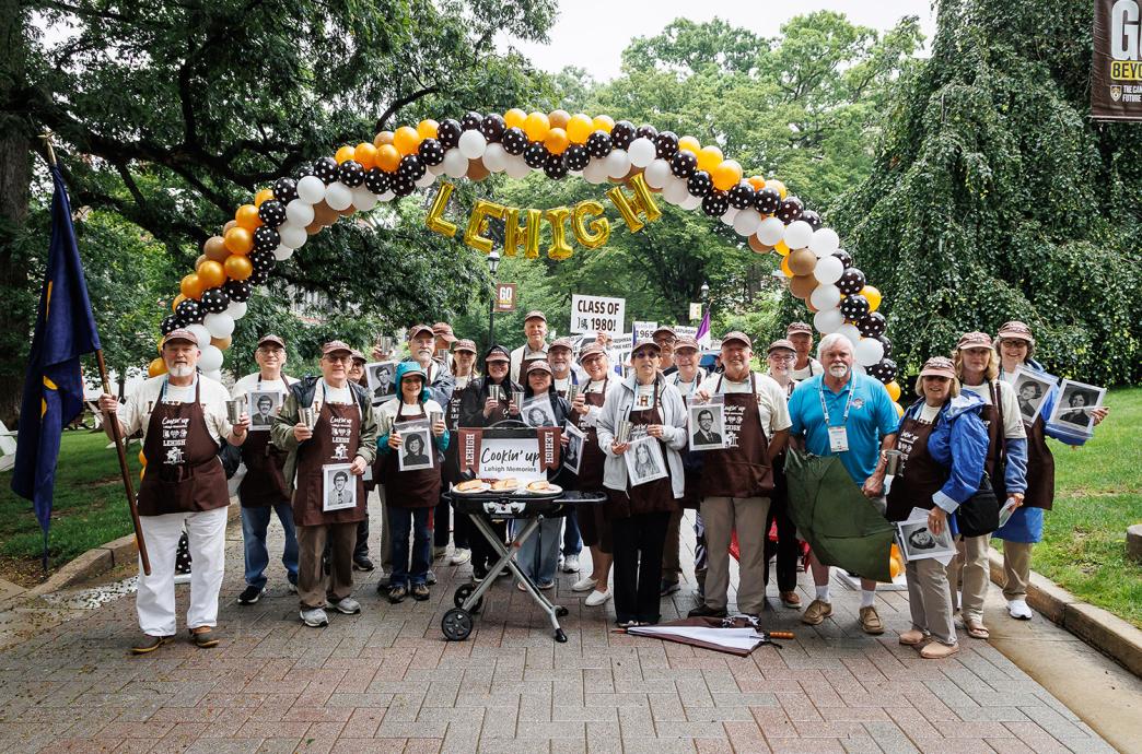 Alumni from class of 1980 wear matching aprons in front of a grill that says “Cookin Up Lehigh Memories”