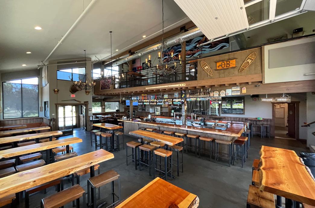 A wide view of the interior of Outlaw Brewing, filled with long tables with live edge-style wood and high-top seating, as well as loft seating above the bar.
