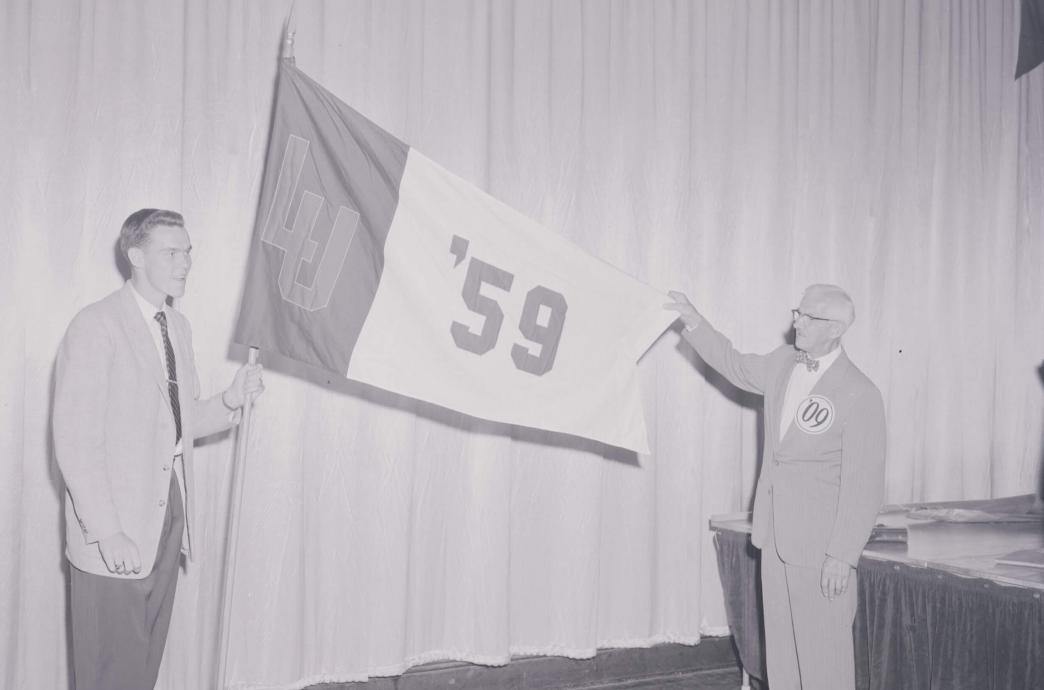 In a black and white photo, a male student standing on a stage holds a large flag that reads “LU ’59” while an older gentleman wearing a large ’09 button holds the flag out to be read.