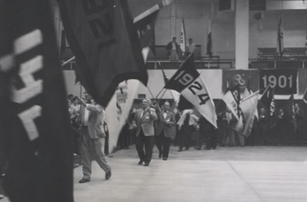 Men wearing suit jackets and ties in a black and white photo parade through an auditorium waving large flags with their class year, one reading 1924.