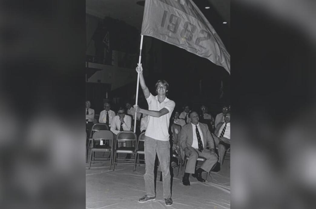 In a black and white photo, a man wearing a white shirt and jeans holds a flag high above his head that says “1982.”