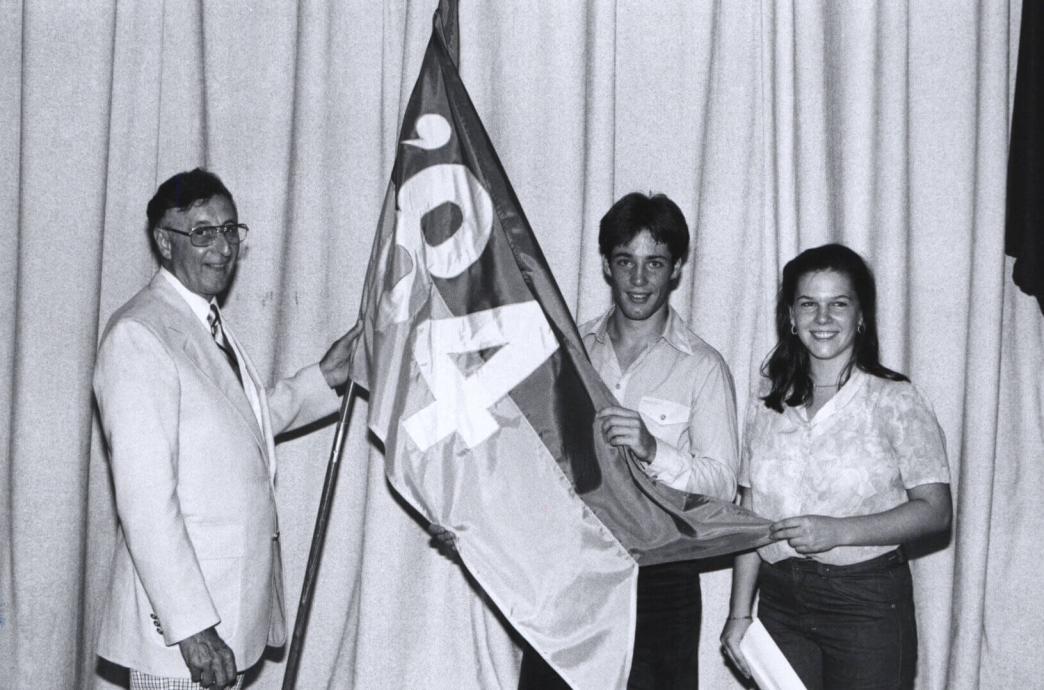 A black and white photo of a man in a light colored suit standing with two students on stage, all holding a large flag together that reads “’84.”