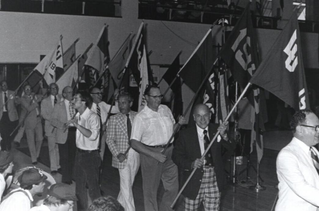 Men line up in a black and white photo, all holding their class flags while parading through an auditorium.