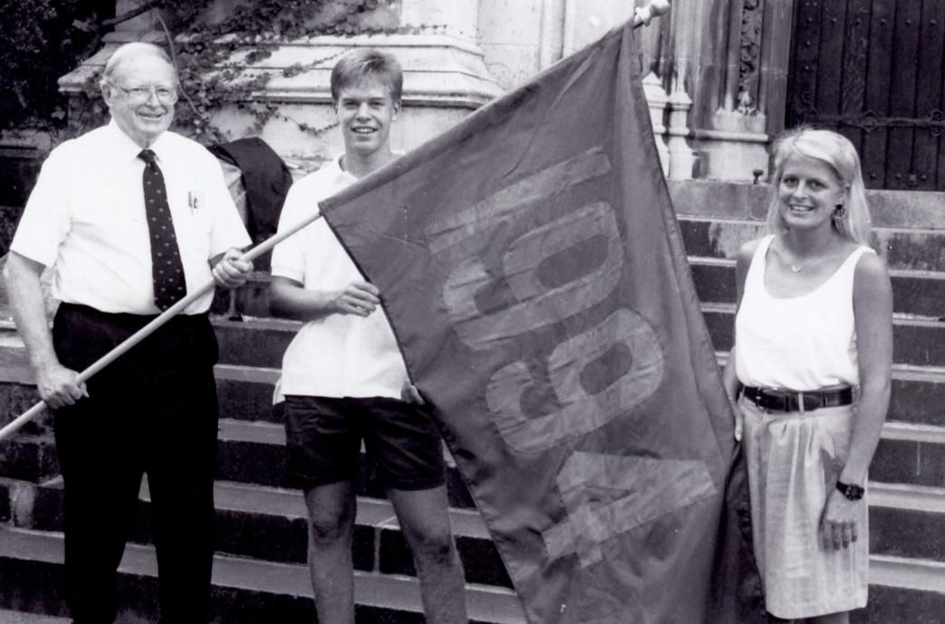 Black and white photo of man wearing a dress shirt, tie and dress pants holding a 1994 flag with two students.