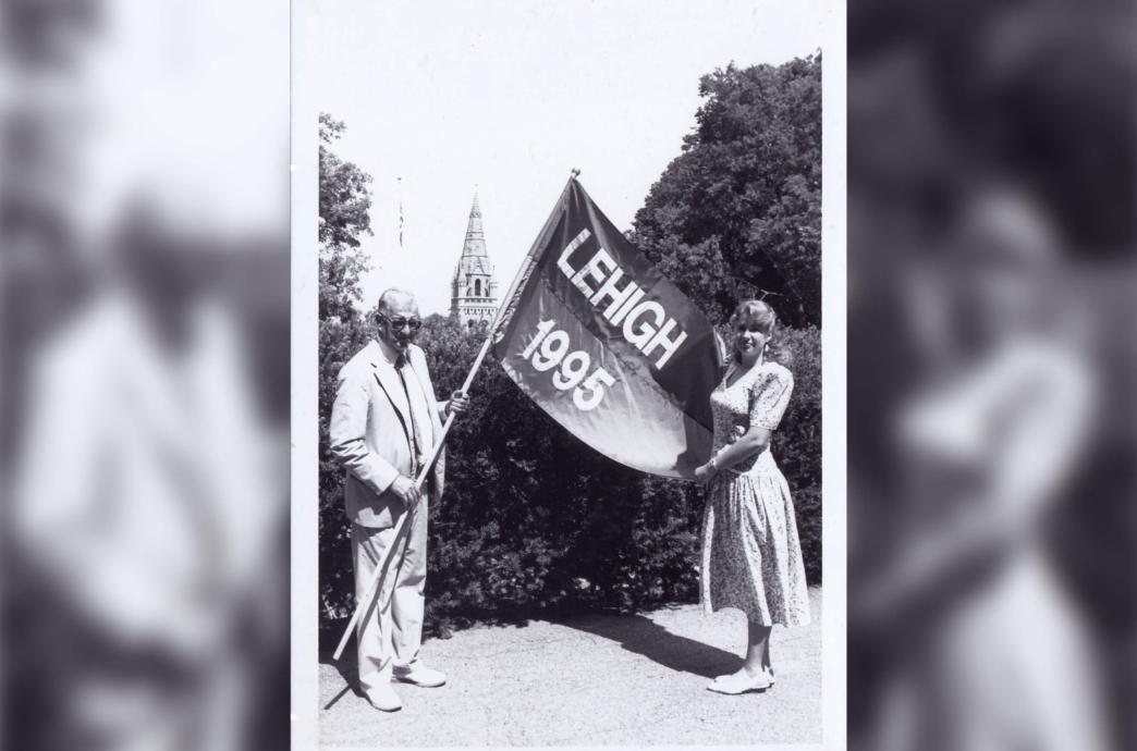 Black and white photo of a man in a light colored suit standing outside and handing a flag that reads “Lehigh 1995” to a young woman in a floral dress.