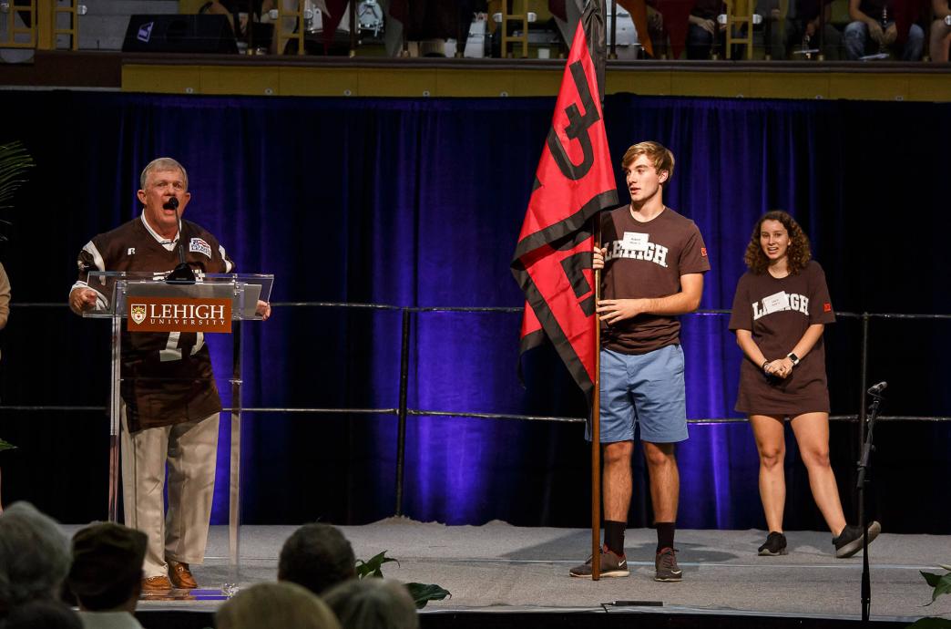 A man wearing a brown Lehigh jersey stands at a podium and yells enthusiastically into the microphone while a male and female student look on holding a red and black class flag.