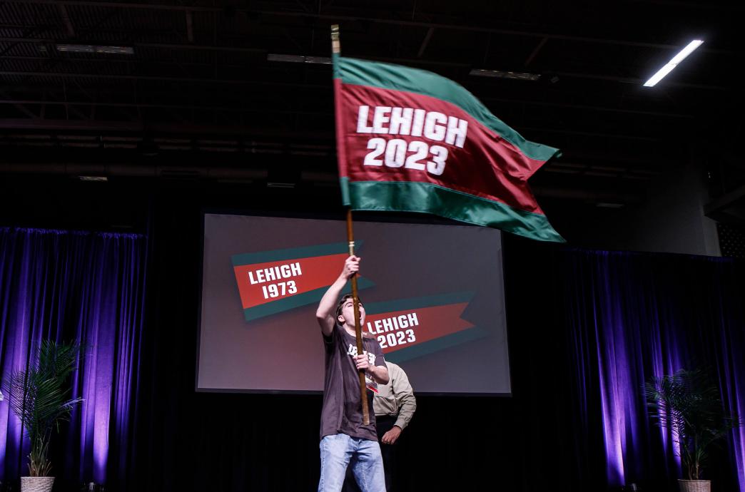 On a dark stage with a large screen behind him, a student waves a green and red flag above his head that reads “Lehigh 2023.”