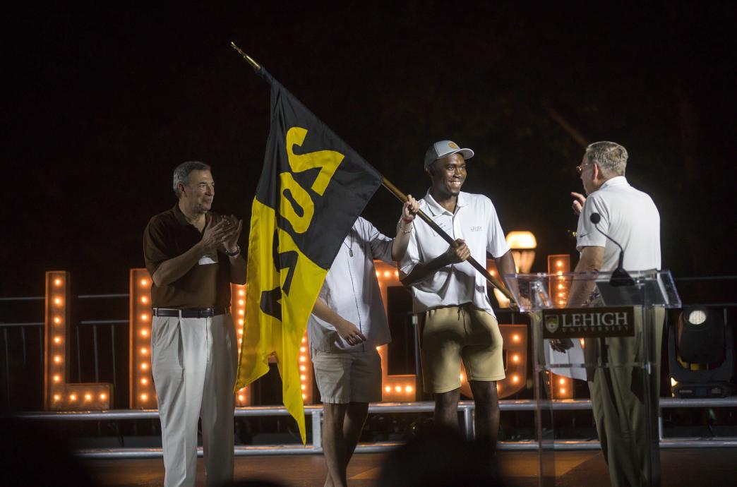 A student wearing a baseball cap with white polo shirt and khaki shorts carries a black and gold flag while standing on stage with 3 other men.