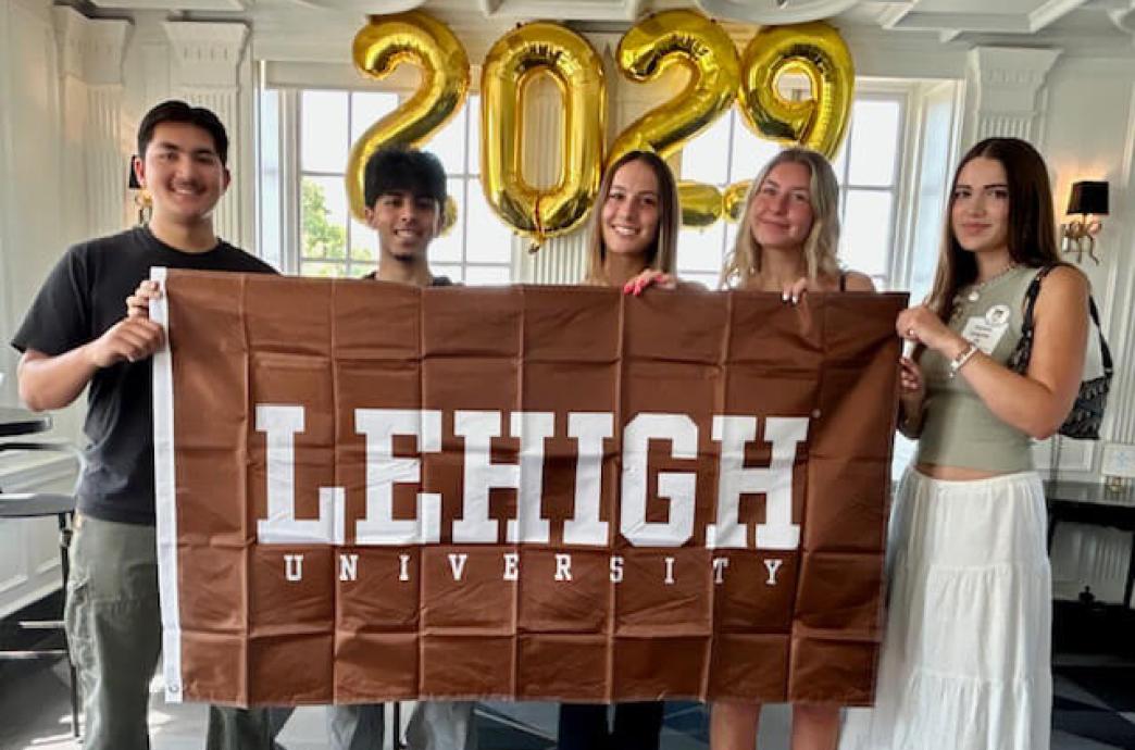 Group of five smiling people holding a Lehigh banner indoors with ”2029” balloons behind them.