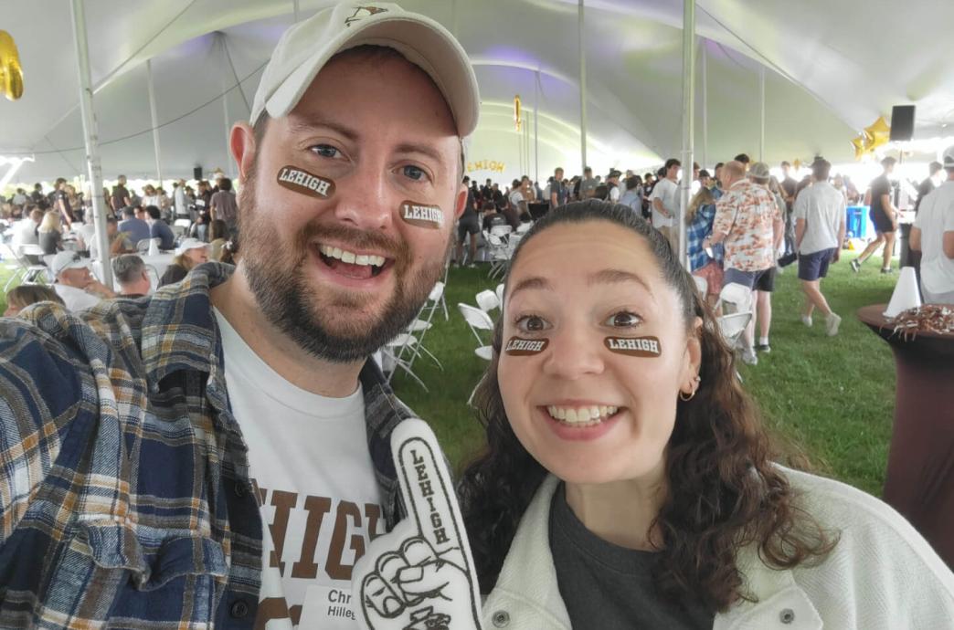 Anderson and Hillegas smile under the alumni tailgate tent at Rivalry holding a foam finger and wearing Lehigh patches under their eyes.