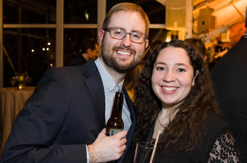 At a semi-formal event, the couple smiles for the camera in a dimly lit room.